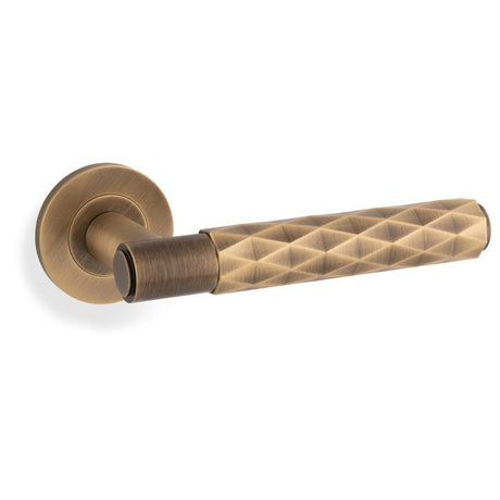 This is an image showing Alexander & Wilks Spitfire Diamond Cut Lever on Round Rose - Antique Brass - aw226-AB available to order from T.H. Wiggans Ironmongery in Kendal, quick delivery and discounted prices.