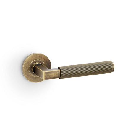 This is an image showing Alexander & Wilks Hurricane Reeded Lever on Round Rose - Antique Brass aw202ab available to order from T.H Wiggans Ironmongery in Kendal, quick delivery and discounted prices.