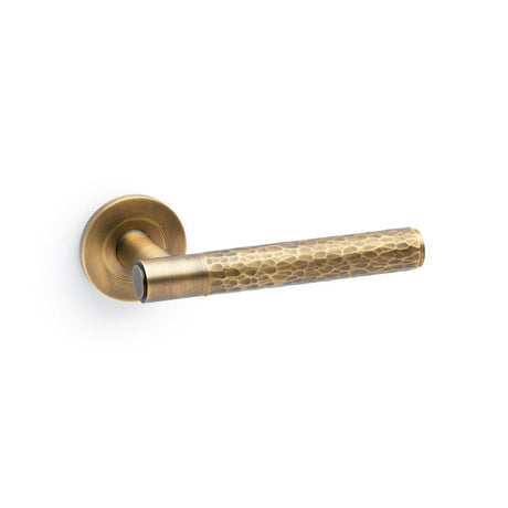This is an image showing Alexander & Wilks Spitfire Hammered Lever on Round Rose - Italian Brass aw223ib available to order from T.H Wiggans Ironmongery in Kendal, quick delivery and discounted prices.