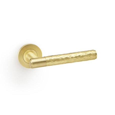 This is an image showing Alexander & Wilks Spitfire Hammered Lever on Round Rose - Satin Brass aw223sb available to order from T.H Wiggans Ironmongery in Kendal, quick delivery and discounted prices.
