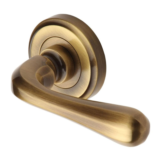 This is an image of a Heritage Brass - Door Handle Lever Latch on Round Rose Charlbury Design Antique Bra, v3020-at that is available to order from T.H Wiggans Ironmongery in Kendal.