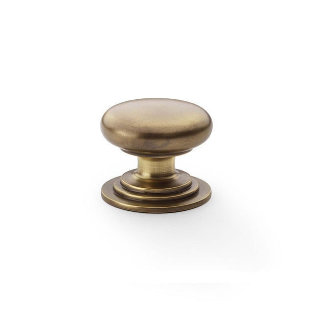 This is an image showing Alexander & Wilks Waltz Round Cupboard Knob on Stepped Rose - Antique Brass - 38mm aw825-38-ab available to order from T.H Wiggans Ironmongery in Kendal, quick delivery and discounted prices.